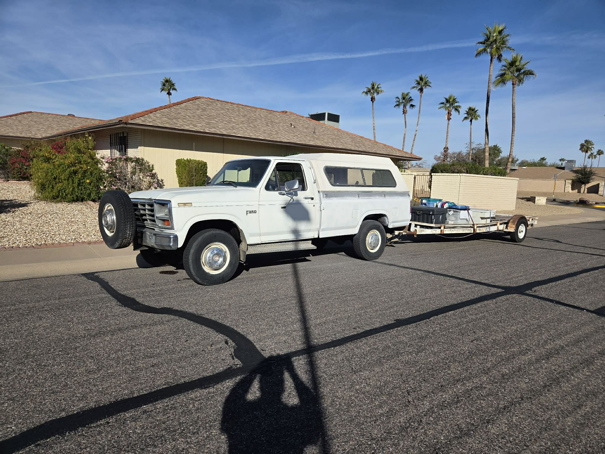 White vintage Ford F-250 with camper shell towing a trailer on a sunny suburban street.